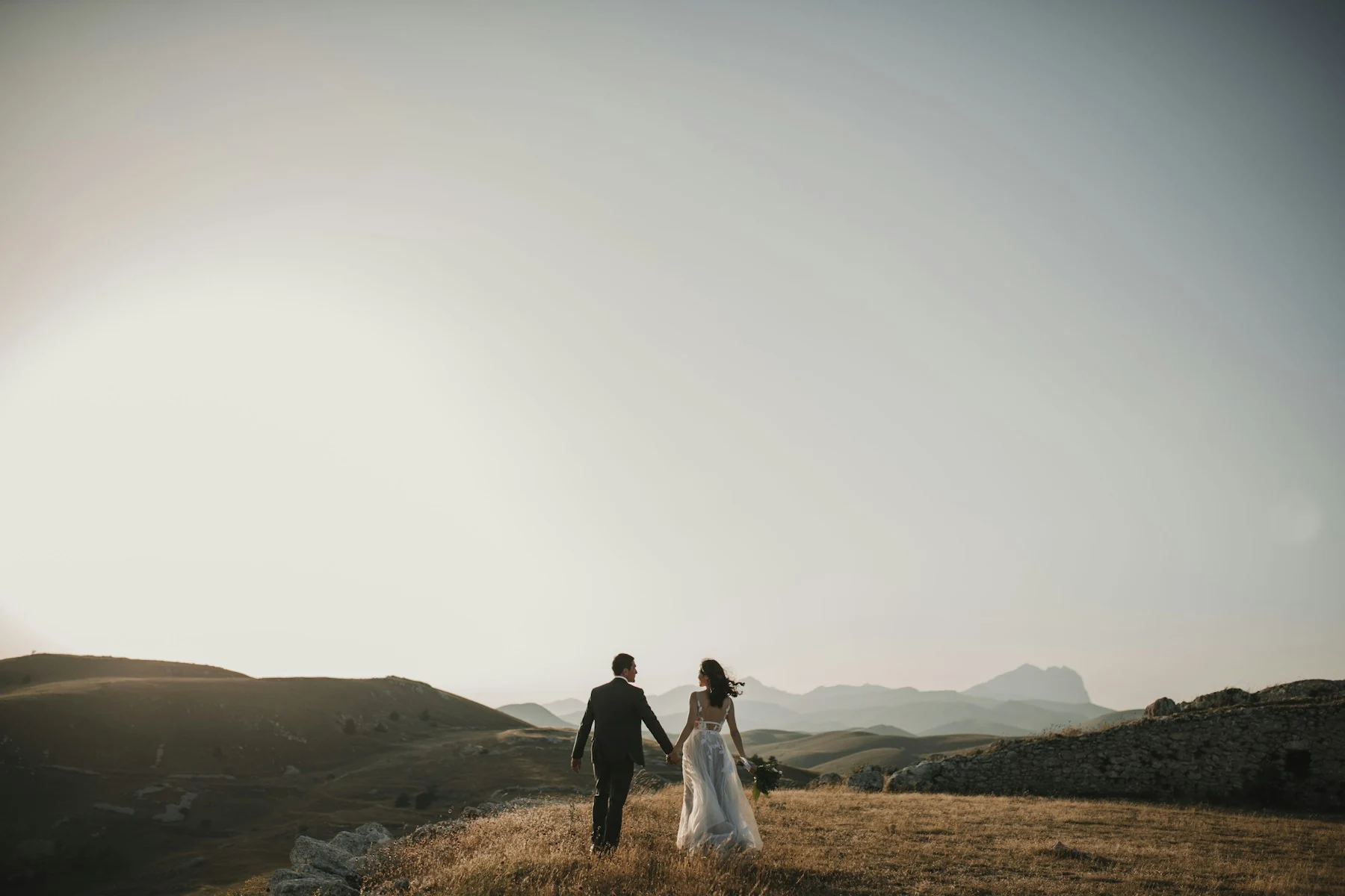 A bride and groom walking hand in hand through a sunlit landscape